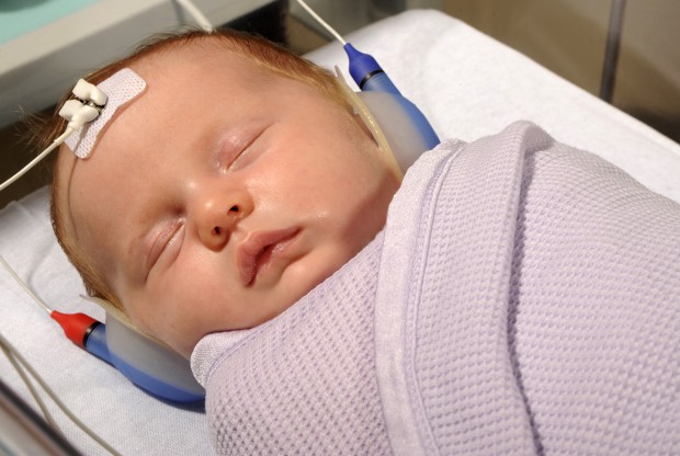 A newborn baby in a cot, with some hearing test equipment on: a headset over the ears and a monitor on the forehead.