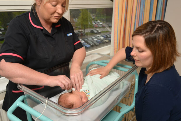 A medical professional performing the newborn hearing screening test on a baby with the mother to one side.