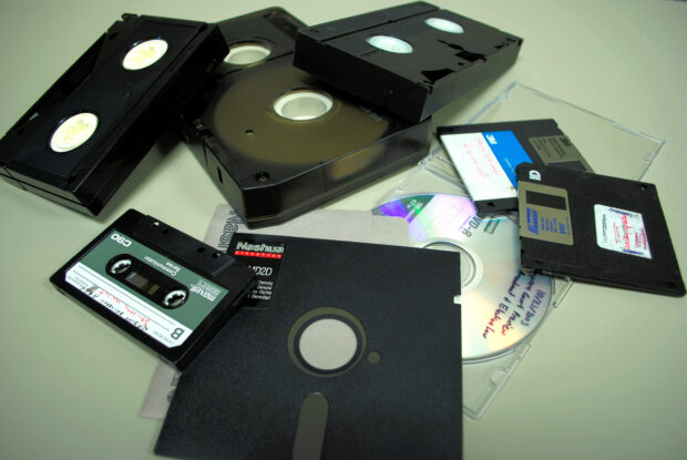 A pile of old media formats: VHS tapes, audio tapes, disk drives, floppy disks.