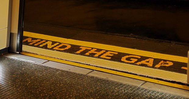 The floor of a tram or tube with the words 'mind the gap' written in bold yellow type.