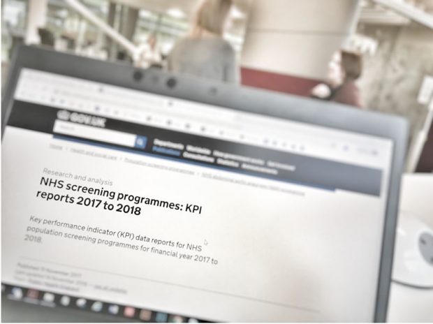 Computer screen screen showing the words 'NHS screening programmes: KPI reports 2017 to 2018' with two women talking in the background.
