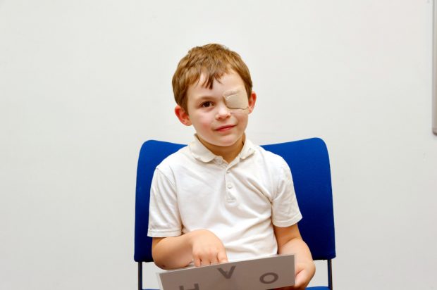 A young boy sitting in chair with an eye patch on and holding a sheet with letters on