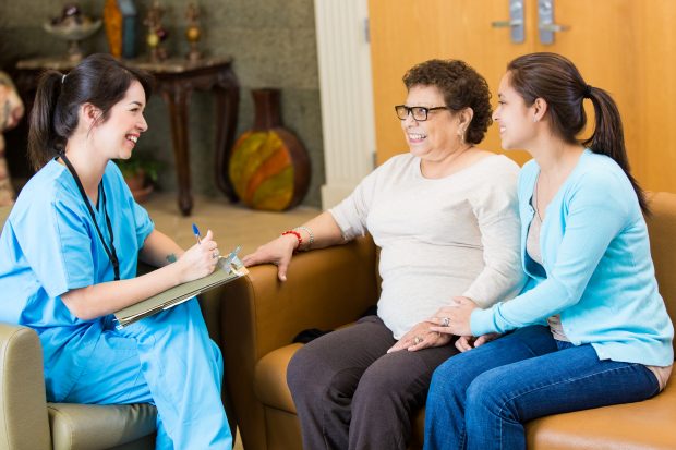 A nurse visiting a female patient and the patient's caregiver or granddaughter. The nurse is visiting the patient at home. The nurse takes notes while talking with the patient.