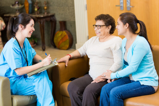 A nurse visiting a female patient and the patient's caregiver or granddaughter. The nurse is visiting the patient at home. The nurse takes notes while talking with the patient.