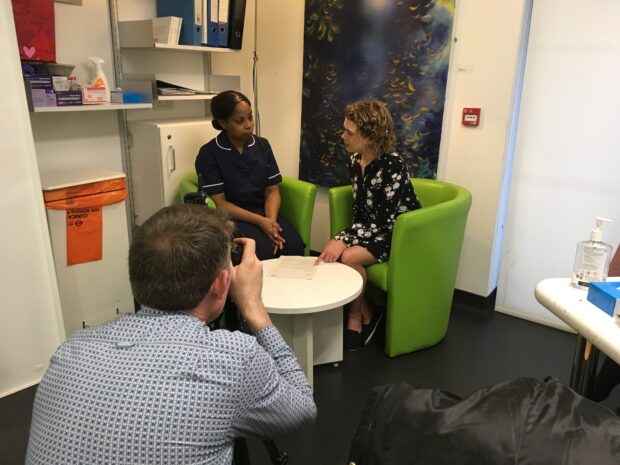 A photographer taking a photo of a woman modelling as a nurse and a woman modelling as a patient at a GP surgery