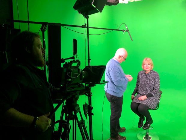 A women sitting on a stool in a film studio with a camera crew setting up around her