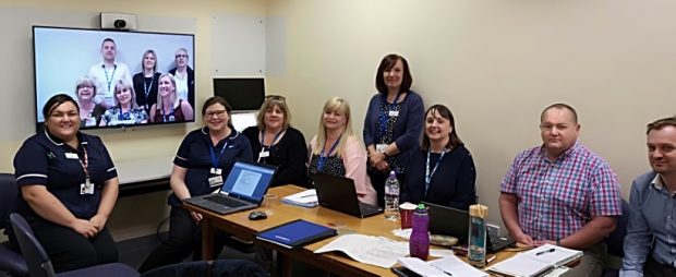 8 people are all sat at a table where they look like they're having a meeting. They are all facing the camera. Several are in nurses uniform.