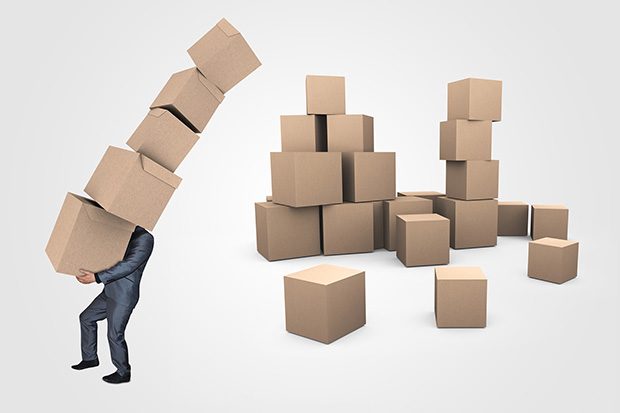 Businessman in a suit struggling under weight of carrying 5 large cardboard boxes. A pile of around 20 more boxes is behind him