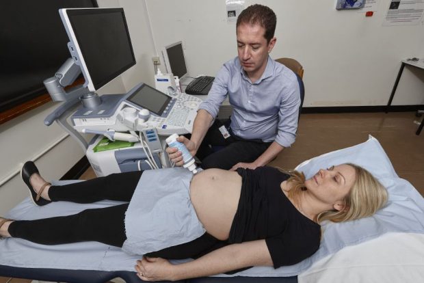 Male sonographer applies gel to pregnant woman's abdomen. She is laid down on a hospital bed.