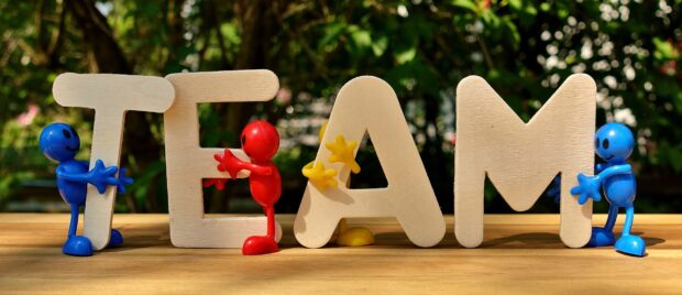 Wooden depictions of the word 'team' are being held on a table by small colourful cartoon people.