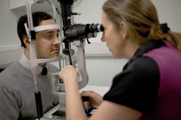 An adult man has his eyes checked by a female member of staff using a piece of equipment called a slit lamp