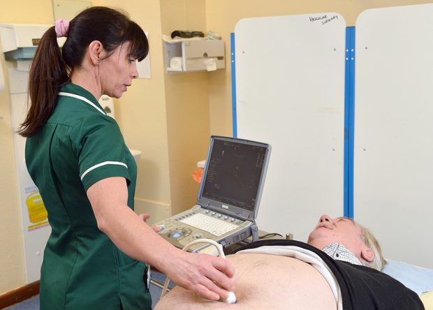 Female AAA screening technician conducts abdominal ultrasound scan on older male lying on couch